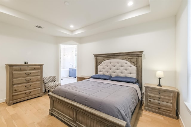 bedroom with light wood-type flooring, ensuite bathroom, and a raised ceiling