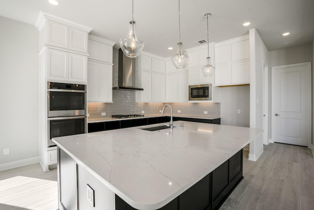 kitchen with stainless steel appliances, a kitchen island with sink, sink, wall chimney range hood, and white cabinets