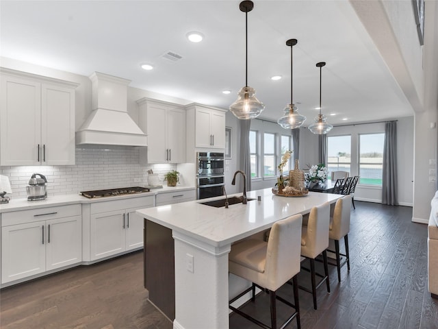 1412 River Point Dr, Friendswood, TX 77546 - kitchen with custom range hood, double oven, backsplash, an island with sink, and dark wood-style flooring