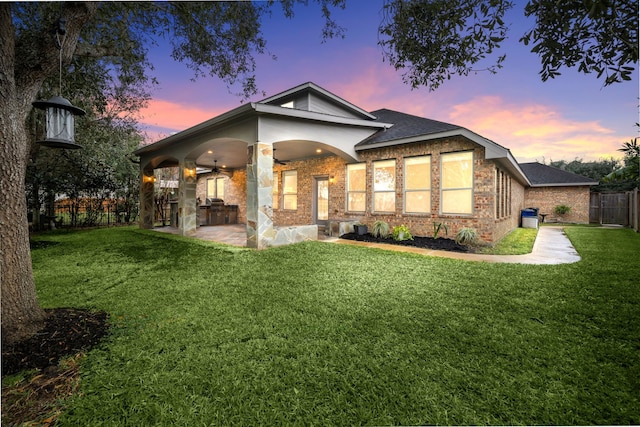 back house at dusk with an outdoor kitchen, ceiling fan, a patio area, and a lawn