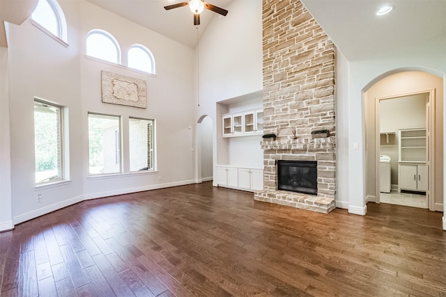 unfurnished living room with ceiling fan, a stone fireplace, a towering ceiling, and dark wood-type flooring