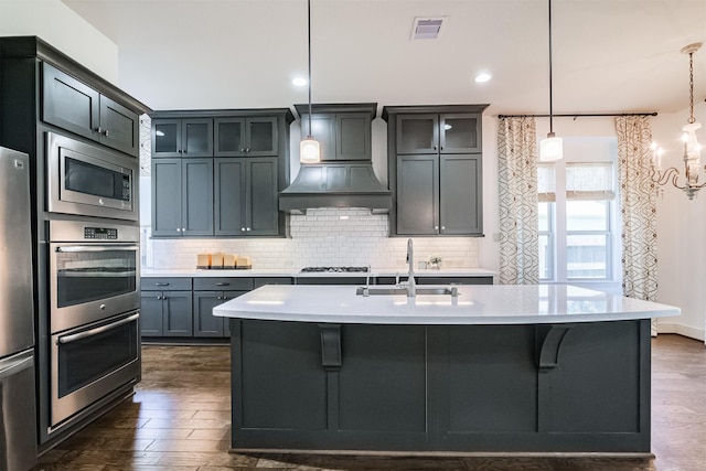 kitchen with custom exhaust hood, a kitchen island with sink, dark wood-type flooring, hanging light fixtures, and appliances with stainless steel finishes