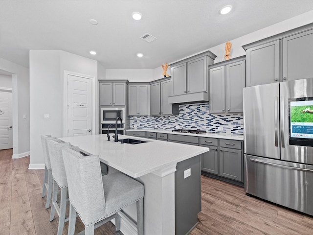 kitchen featuring a center island with sink, stainless steel appliances, tasteful backsplash, gray cabinets, and a sink