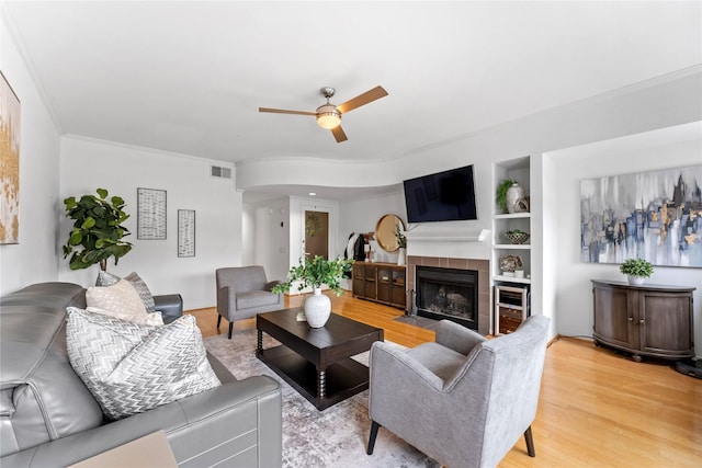 living room with built in shelves, crown molding, a tile fireplace, ceiling fan, and light hardwood / wood-style floors