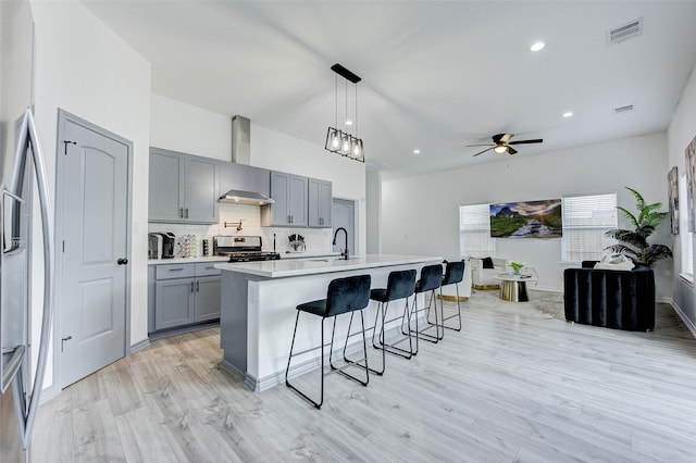 kitchen featuring gray cabinetry, a center island with sink, appliances with stainless steel finishes, pendant lighting, and wall chimney range hood