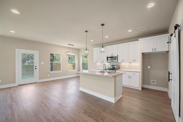 kitchen with light stone countertops, stainless steel appliances, a barn door, a center island with sink, and white cabinets