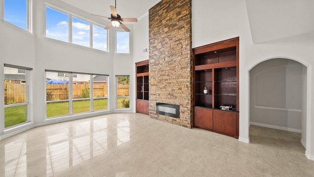 unfurnished living room featuring built in shelves, ceiling fan, a stone fireplace, plenty of natural light, and a towering ceiling