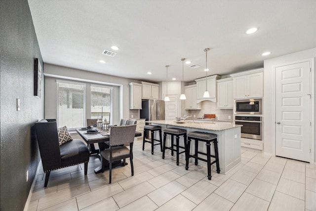 kitchen featuring a center island with sink, decorative backsplash, appliances with stainless steel finishes, decorative light fixtures, and white cabinetry