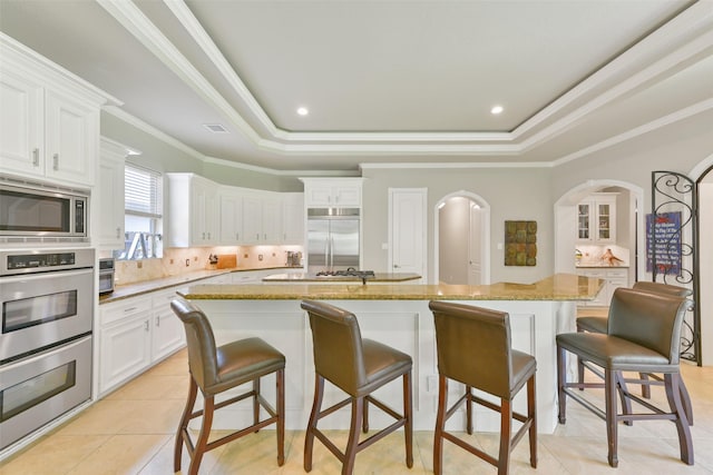 kitchen featuring a tray ceiling, built in appliances, white cabinets, and an island with sink