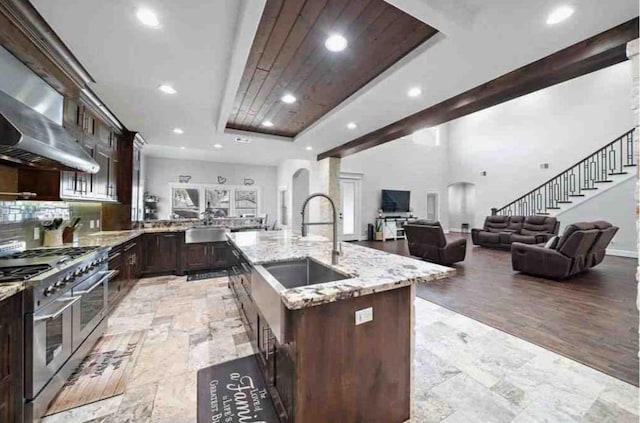 kitchen with light stone counters, a kitchen island with sink, wall chimney range hood, sink, and double oven range