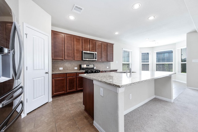 kitchen featuring backsplash, a kitchen island with sink, light tile patterned floors, appliances with stainless steel finishes, and light stone counters