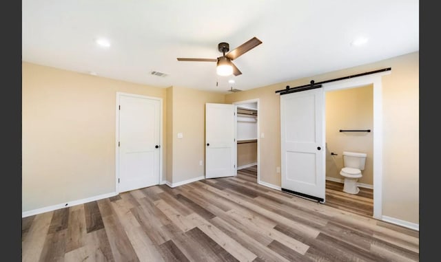 unfurnished bedroom featuring connected bathroom, ceiling fan, a barn door, light hardwood / wood-style flooring, and a closet