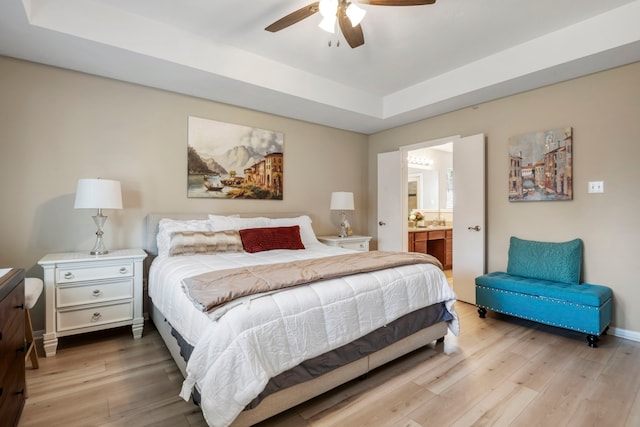 bedroom with ceiling fan, a raised ceiling, light wood-type flooring, and ensuite bath