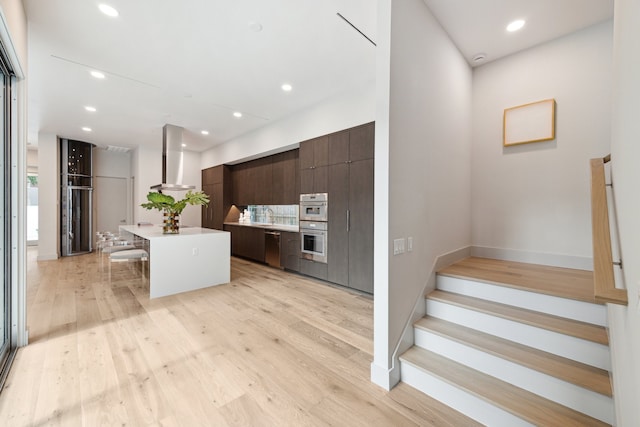 kitchen featuring a center island, wall chimney range hood, a kitchen breakfast bar, light hardwood / wood-style floors, and dark brown cabinets