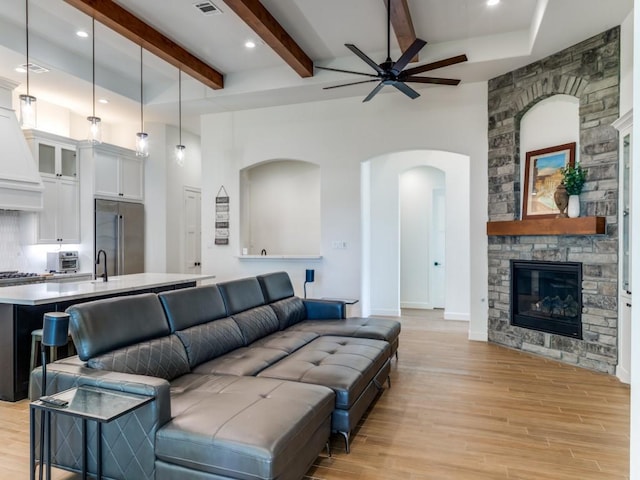 living room featuring beamed ceiling, light wood-type flooring, a stone fireplace, and ceiling fan