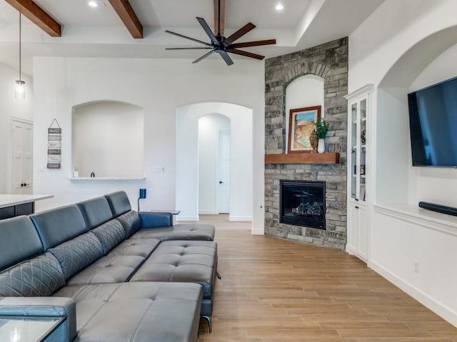 living room with a stone fireplace, ceiling fan, beamed ceiling, and light wood-type flooring