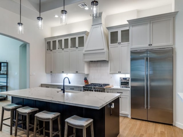 kitchen featuring light stone counters, stainless steel built in fridge, decorative light fixtures, and a center island with sink