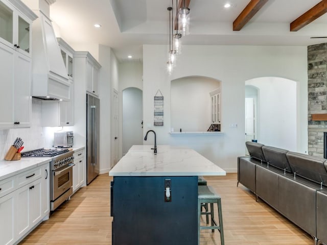 kitchen featuring premium appliances, beamed ceiling, premium range hood, decorative light fixtures, and a kitchen island with sink