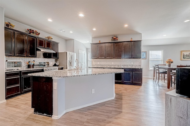 kitchen featuring light stone countertops, appliances with stainless steel finishes, decorative backsplash, dark brown cabinets, and a kitchen island with sink