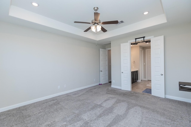 unfurnished bedroom featuring ceiling fan, light colored carpet, a raised ceiling, and ensuite bath