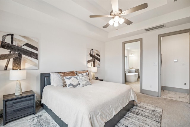 bedroom with a raised ceiling, light colored carpet, ensuite bath, and ceiling fan