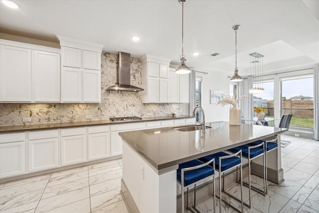 kitchen featuring pendant lighting, white cabinets, sink, wall chimney exhaust hood, and an island with sink