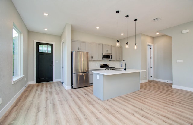 kitchen with gray cabinetry, hanging light fixtures, stainless steel appliances, an island with sink, and light hardwood / wood-style floors