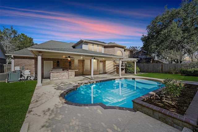 pool at dusk with ceiling fan, a yard, a patio, and an outdoor kitchen