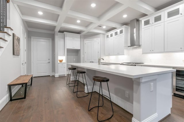 kitchen featuring coffered ceiling, wall chimney range hood, beamed ceiling, white cabinets, and an island with sink