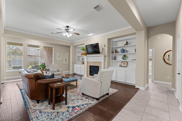 living room with built in shelves, ornamental molding, a tile fireplace, ceiling fan, and light hardwood / wood-style floors