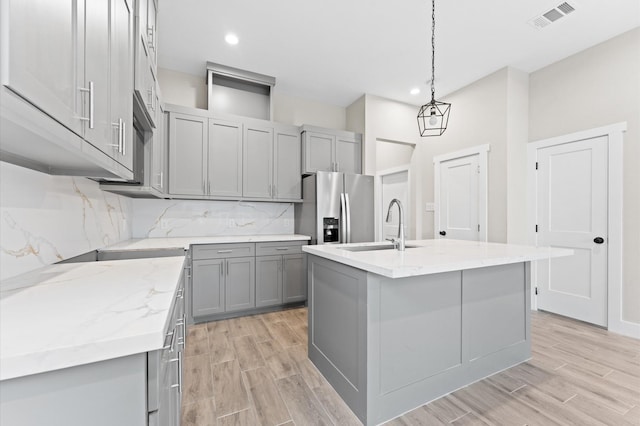 kitchen with pendant lighting, sink, stainless steel refrigerator with ice dispenser, gray cabinets, and an island with sink