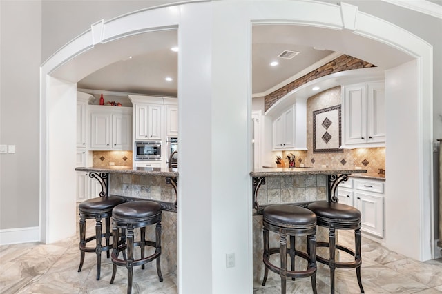 kitchen featuring stone counters, white cabinetry, stainless steel microwave, crown molding, and a breakfast bar