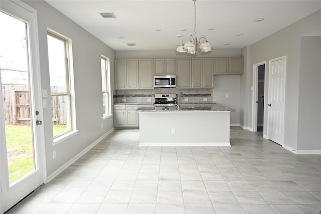 kitchen featuring stove, backsplash, light stone counters, a center island with sink, and decorative light fixtures