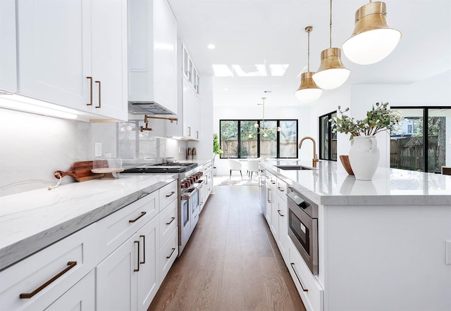 kitchen with sink, a center island with sink, range with two ovens, white cabinetry, and hanging light fixtures