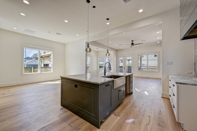 kitchen featuring hanging light fixtures, ceiling fan, light stone countertops, an island with sink, and white cabinetry