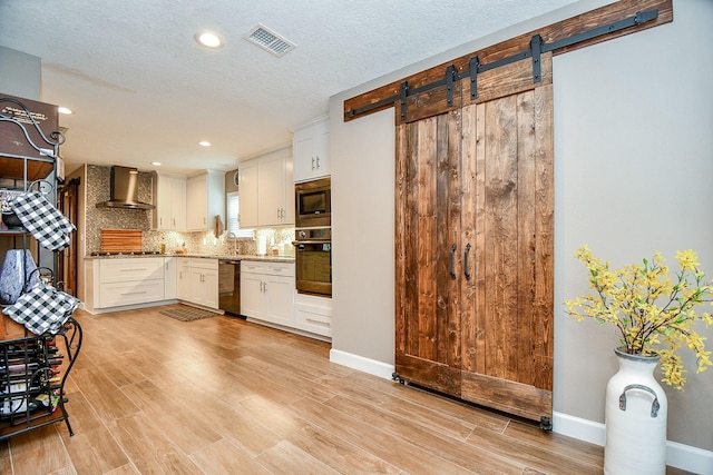 kitchen with a barn door, wall chimney range hood, white cabinets, and black appliances