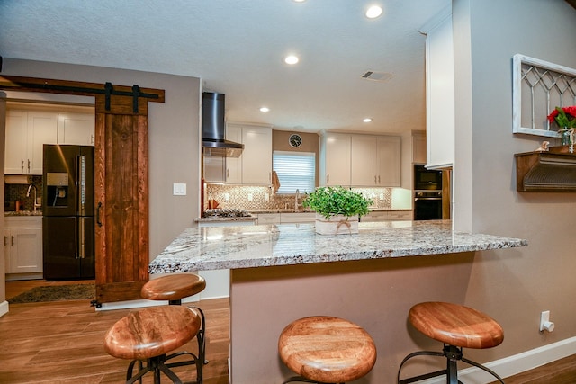 kitchen with black appliances, a barn door, wall chimney exhaust hood, and a kitchen bar