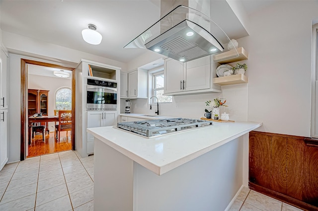 kitchen featuring white cabinetry, kitchen peninsula, island range hood, light tile patterned flooring, and appliances with stainless steel finishes