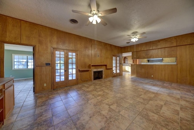 unfurnished living room featuring wood walls, french doors, ceiling fan, a textured ceiling, and a tiled fireplace