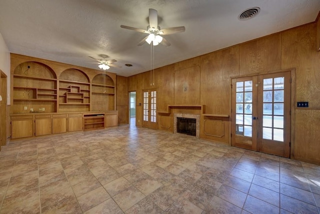 unfurnished living room with a tile fireplace, french doors, wooden walls, ceiling fan, and built in shelves