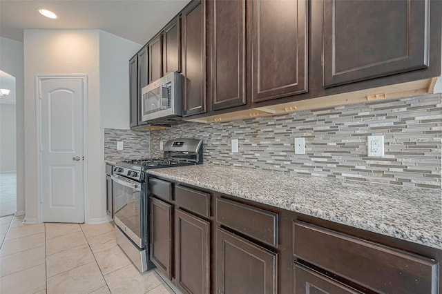 kitchen with appliances with stainless steel finishes, backsplash, light stone counters, dark brown cabinets, and light tile patterned flooring