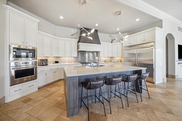 kitchen with pendant lighting, wall chimney range hood, white cabinetry, built in appliances, and an island with sink