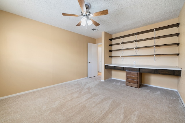 unfurnished office with ceiling fan, light colored carpet, built in desk, and a textured ceiling