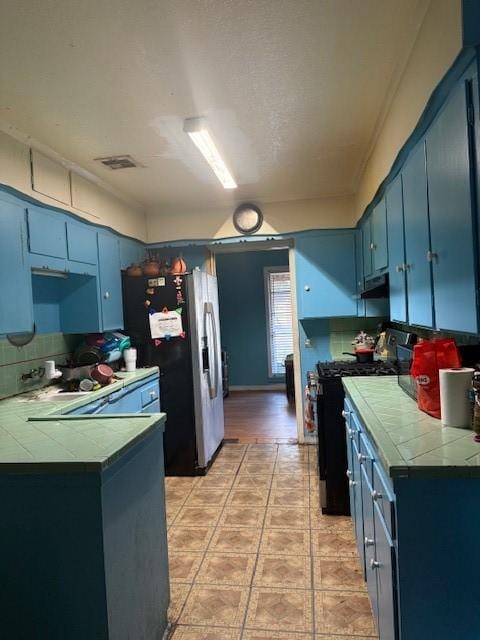 kitchen with blue cabinetry, tile countertops, stainless steel fridge, black gas stove, and kitchen peninsula