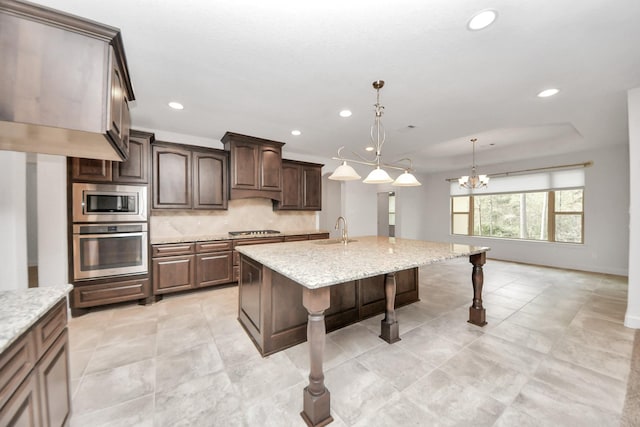 kitchen featuring stainless steel appliances, light stone counters, hanging light fixtures, a chandelier, and a kitchen island with sink