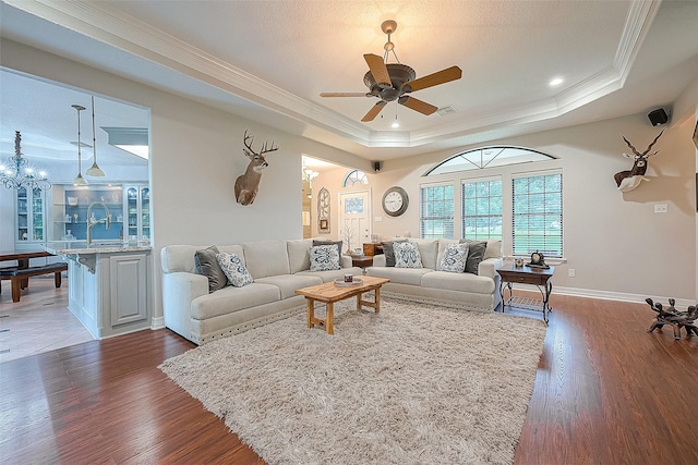 living room featuring sink, dark wood-type flooring, ornamental molding, ceiling fan with notable chandelier, and a raised ceiling