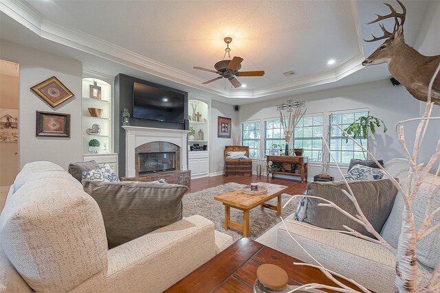 living room with wood-type flooring, ornamental molding, ceiling fan, a raised ceiling, and built in shelves