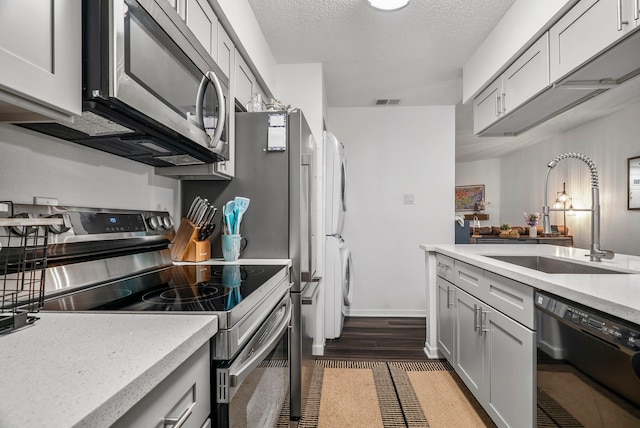 kitchen with sink, gray cabinets, a textured ceiling, appliances with stainless steel finishes, and stacked washer / dryer