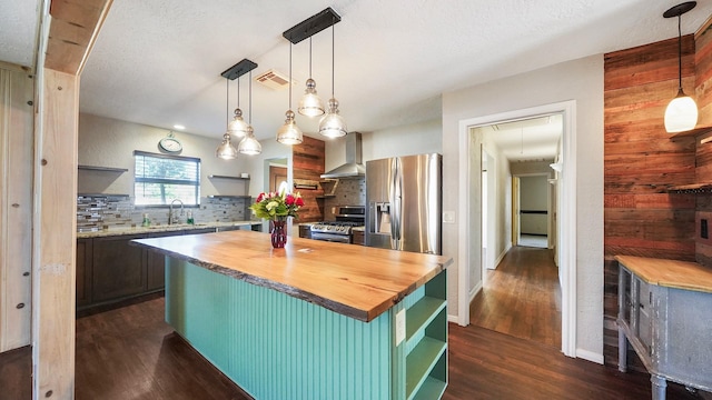 kitchen featuring stainless steel appliances, wall chimney range hood, butcher block countertops, pendant lighting, and a kitchen island