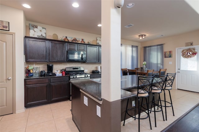 kitchen with stainless steel range with gas cooktop, dark stone countertops, light tile patterned flooring, dark brown cabinetry, and a breakfast bar area